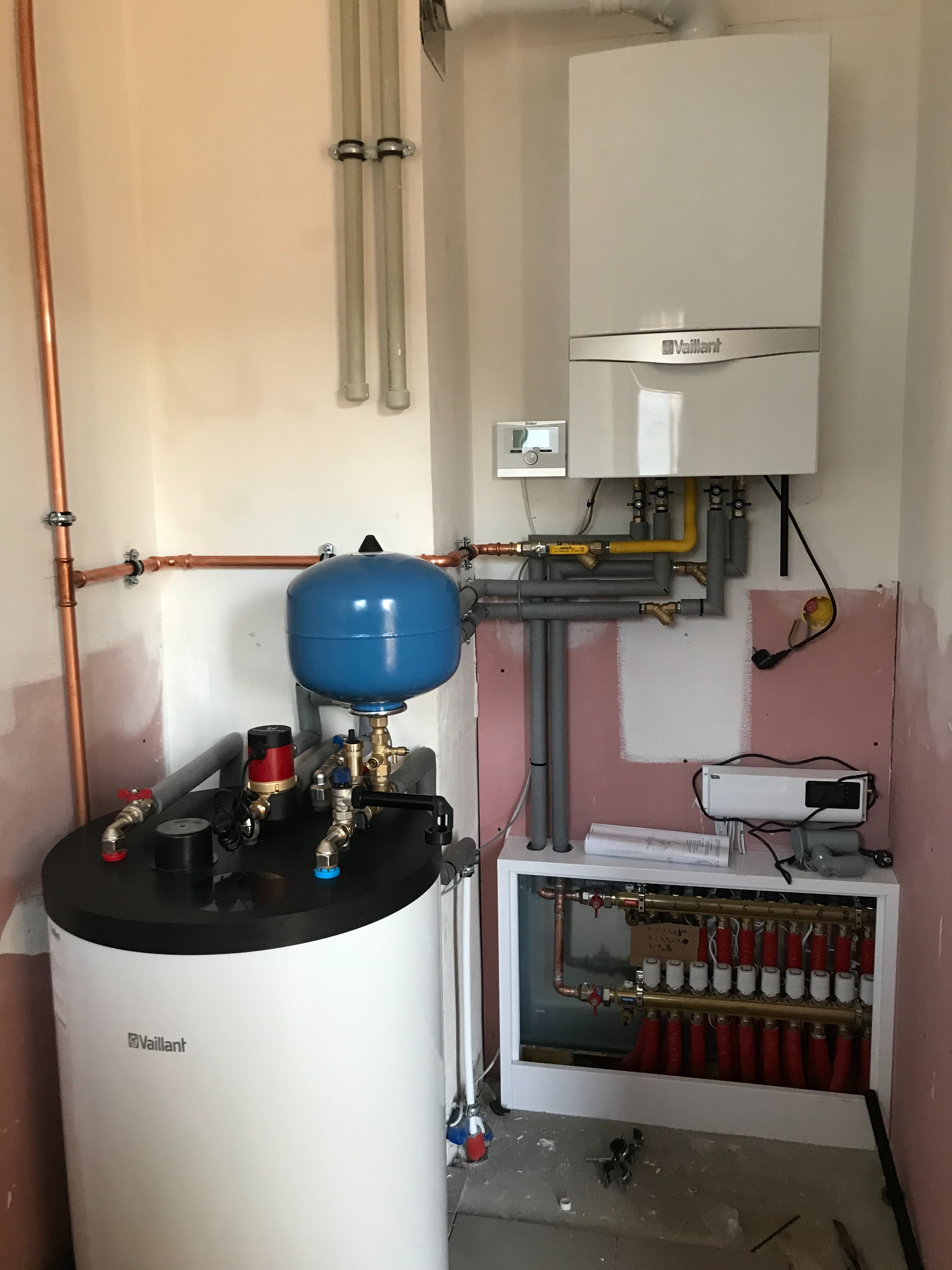 Biały piec gazowy Vaillant, zbiornik z niebieskim naczyniem przeponowym, rozdzielacz ogrzewania podłogowego z czerwonymi pętlami, miedziane rury instalacji w surowym pomieszczeniu.