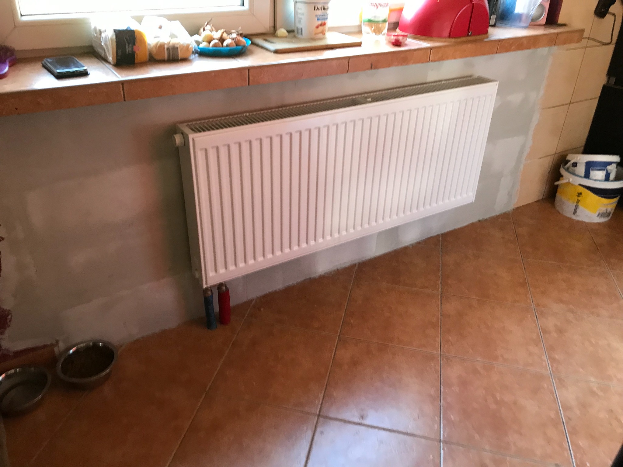 Biały, panelowy grzejnik centralnego ogrzewania zamontowany na ścianie, z widocznymi rurami doprowadzającymi wodę, na tle terakotowej podłogi i ściany w trakcie wykańczania.