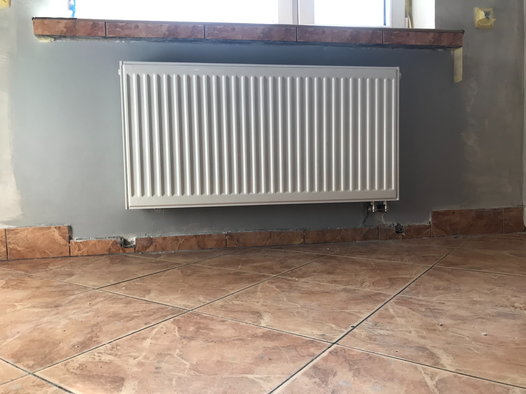 Biały, nowy grzejnik panelowy zamontowany na szarej ścianie w trakcie remontu, widoczne płytki podłogowe i fragment parapetu z płytek, surowe wykończenie ściany.
