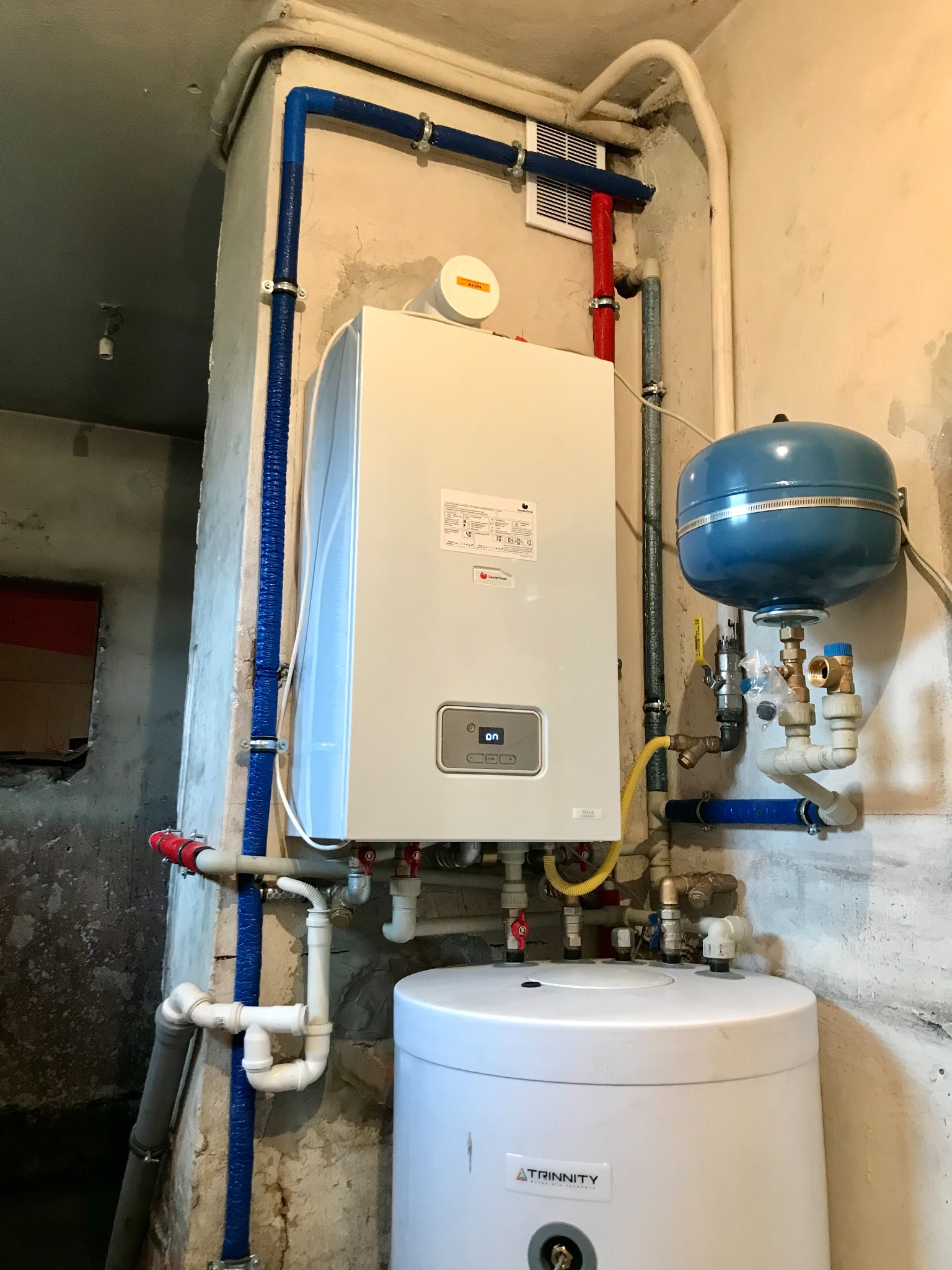 Biały piec gazowy marki De Dietrich Thelia Condens z niebieskim naczyniem przeponowym i białym zasobnikiem Trinnity, zamontowany na ścianie z widocznymi rurami w niebieskiej karbowanej osłonie.