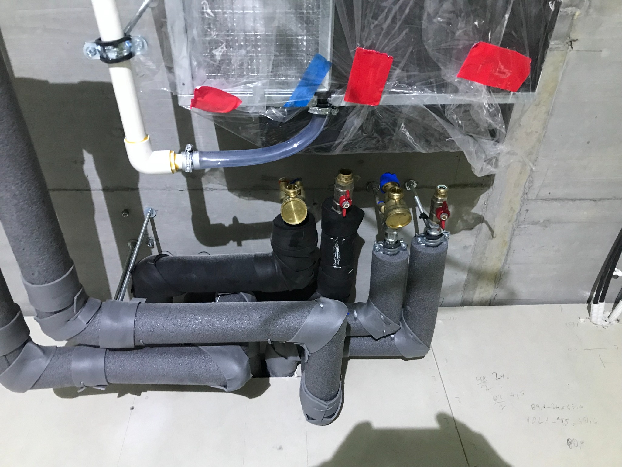 Szare, zaizolowane rury hydrauliczne z zaworami i elementami montażowymi, instalacja w trakcie realizacji, widoczne notatki na podłodze.