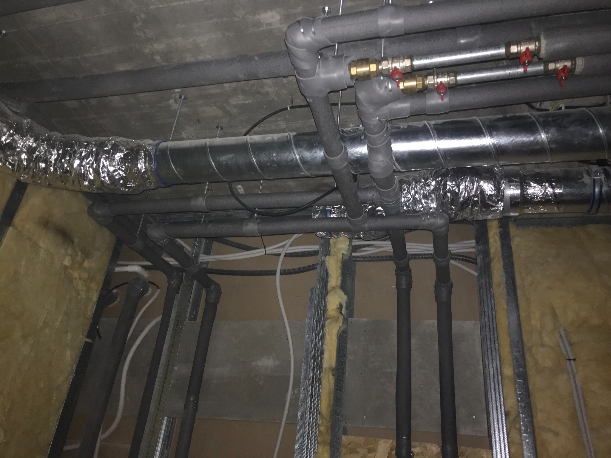 Różne rury instalacyjne pod sufitem, w tym rury izolowane termicznie, rura wentylacyjna w folii aluminiowej i rury z zaworami, widoczne elementy konstrukcyjne ściany z izolacją.