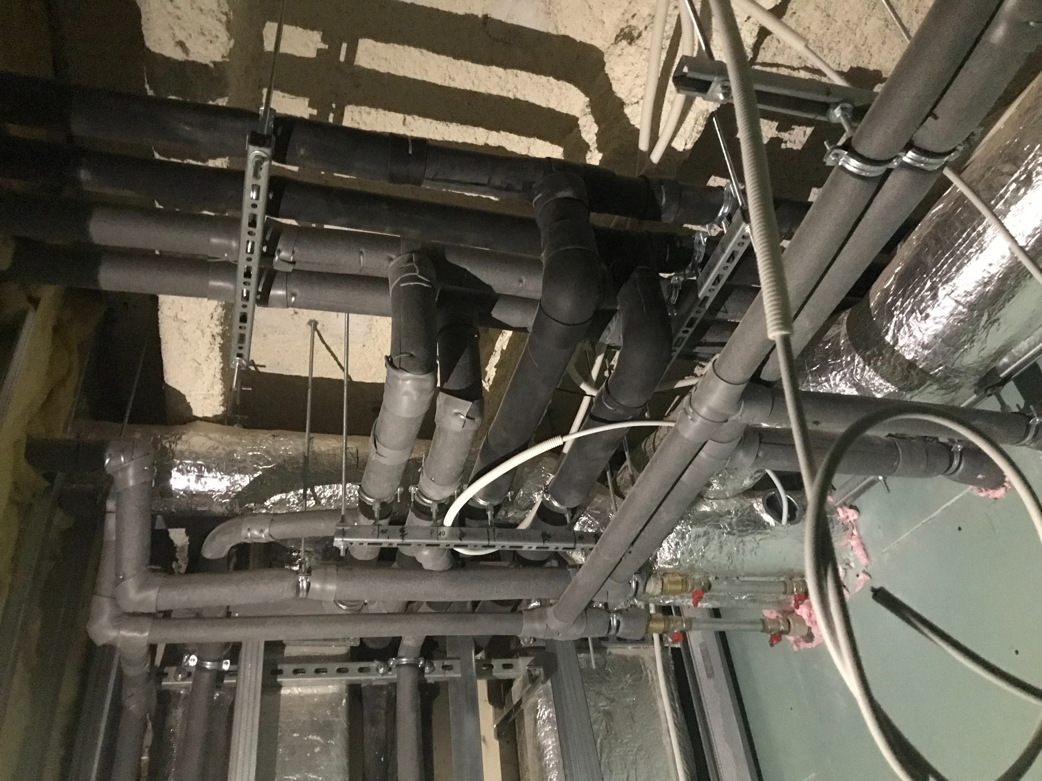 Skomplikowany układ rur instalacyjnych pod sufitem piwnicy, widoczne rury izolowane termicznie, metalowe wsporniki i fragmenty instalacji z zaworami, oświetlenie halogenowe.