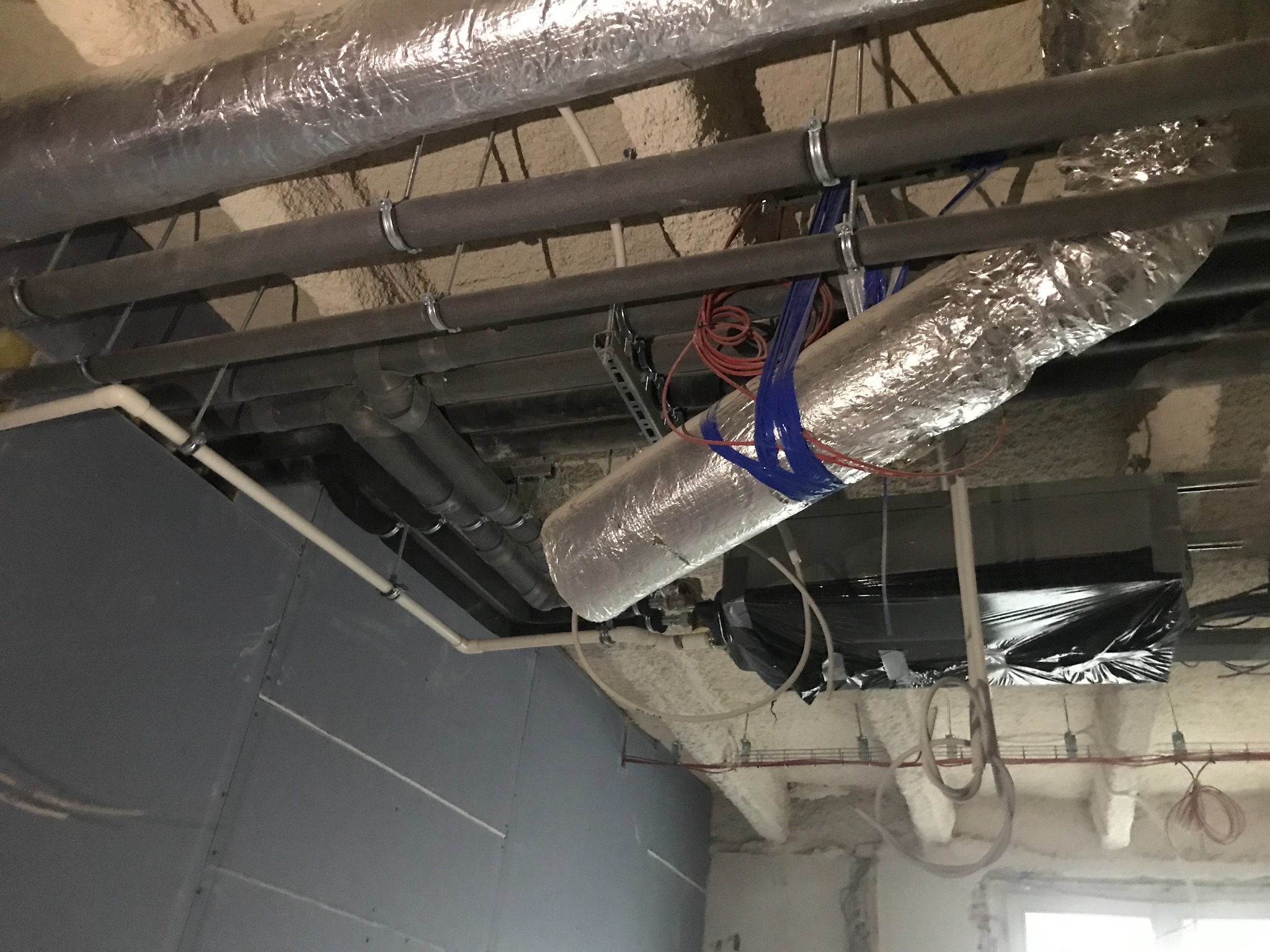 Widok na sufit w trakcie prac instalacyjnych: rury izolowane folią aluminiową, rury PCV, przewody elektryczne, elementy wentylacji, oraz częściowo wykończona ściana z płyt gipsowo-kartonowych.