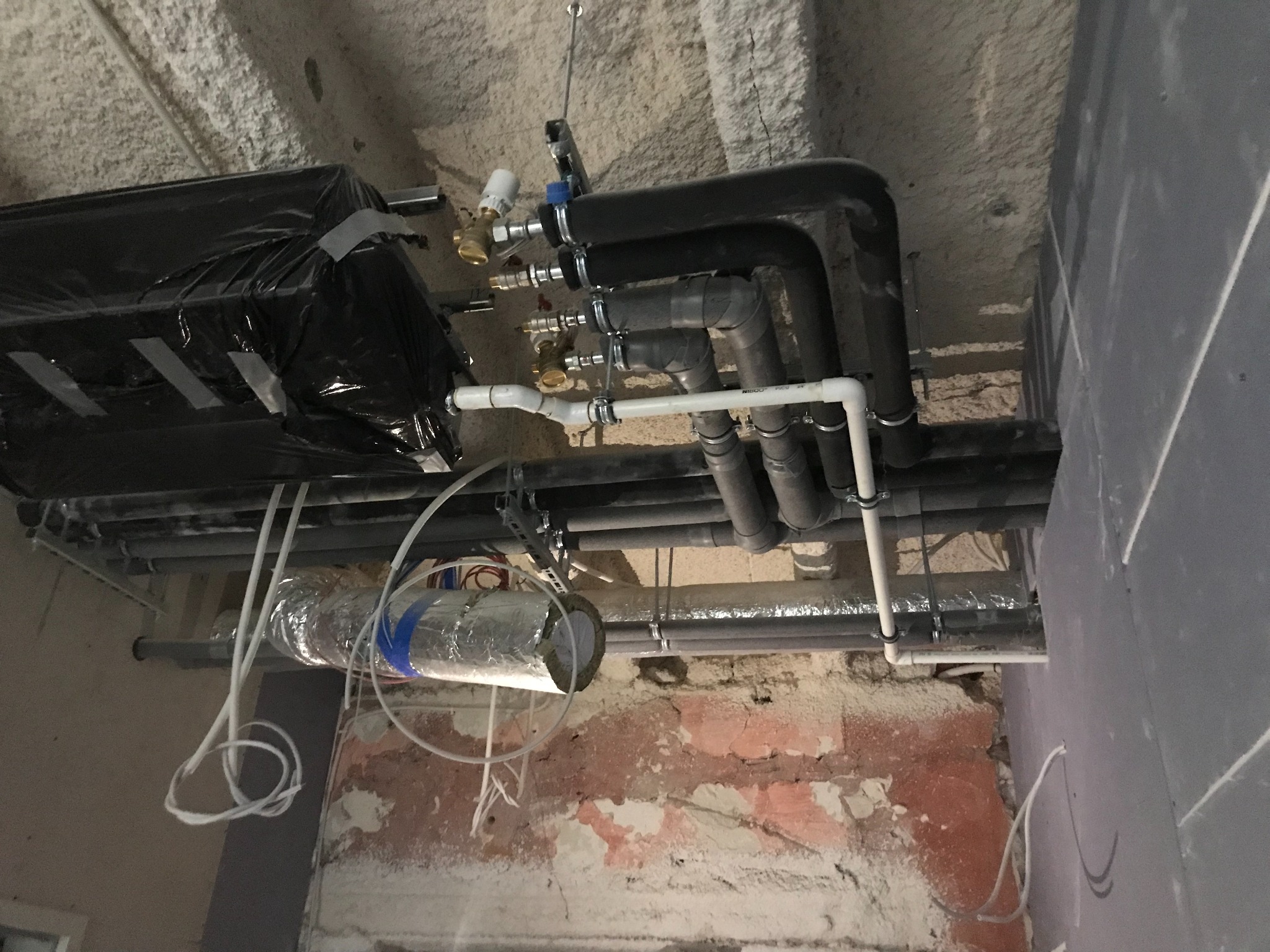 Widok instalacji hydraulicznej i grzewczej pod sufitem w trakcie montażu: rury czarne i białe, zawory, izolacja termiczna owinięta folią aluminiową i czarną folią, surowy betonowy sufit i fragment...