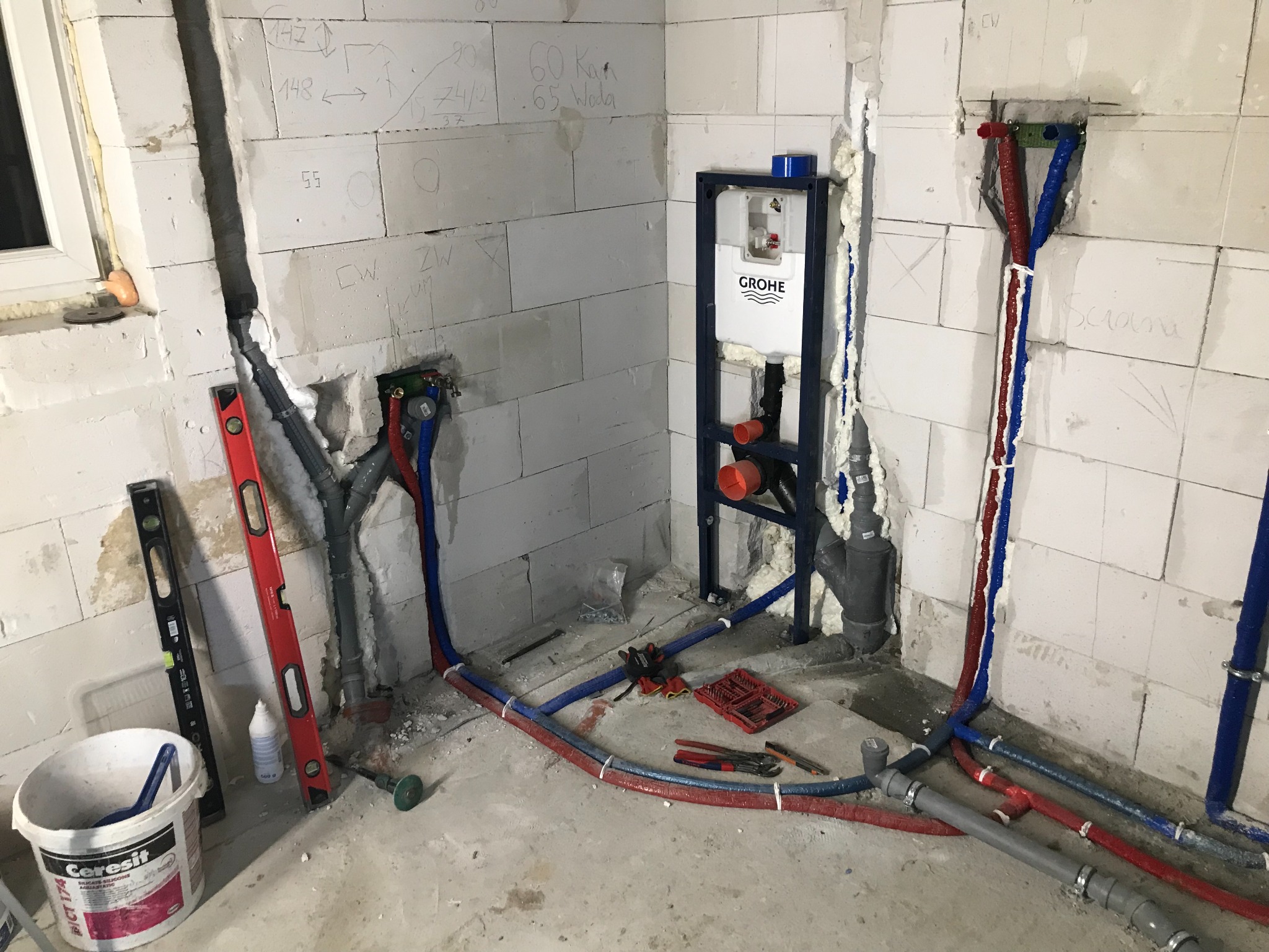 Montaż podtynkowego stelaża WC Grohe z instalacją hydrauliczną w surowym stanie, widoczne rury PEX w izolacji czerwonej i niebieskiej, poziomice, wiadro z zaprawą Ceresit oraz narzędzia...