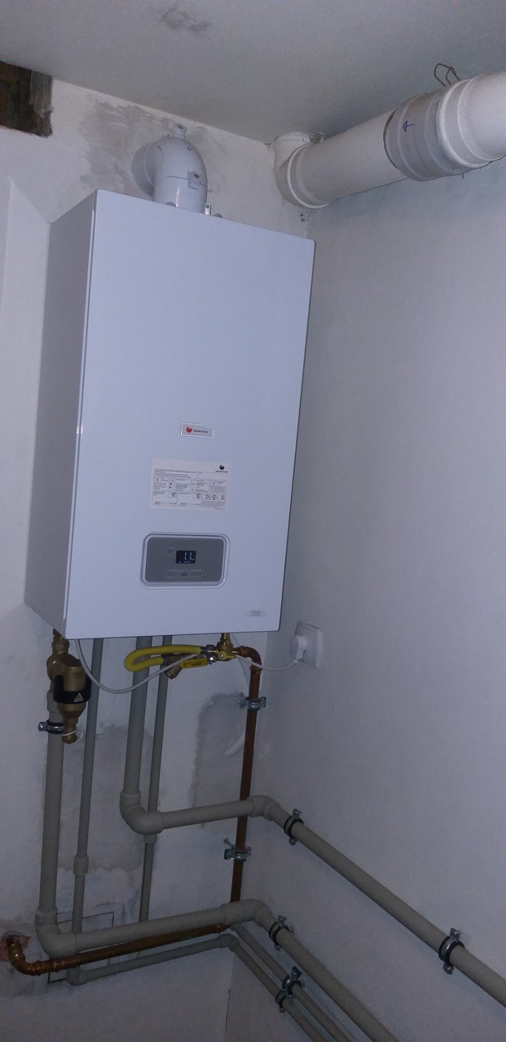 Nowoczesny, biały piec gazowy marki De Dietrich, zamontowany na ścianie z widocznymi rurami gazowymi i wodnymi, w surowym wnętrzu z nieotynkowaną ścianą.