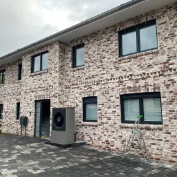 Dwupiętrowy dom z cegły klinkierowej z czarnymi oknami PCV, zewnętrzną pompą ciepła Buderus i drabina oparta o ścianę.