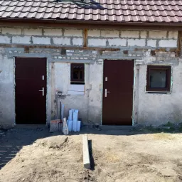 Budynek w trakcie remontu z dwoma nowymi, brązowymi drzwiami wejściowymi, jednym małym oknem i oknem dachowym, widoczne narzędzia budowlane i materiały, teren przed budynkiem w stanie surowym.