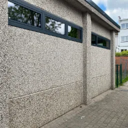 Szare okna w budynku z elewacją z kamyczków; w szybach odbijają się drzewa.