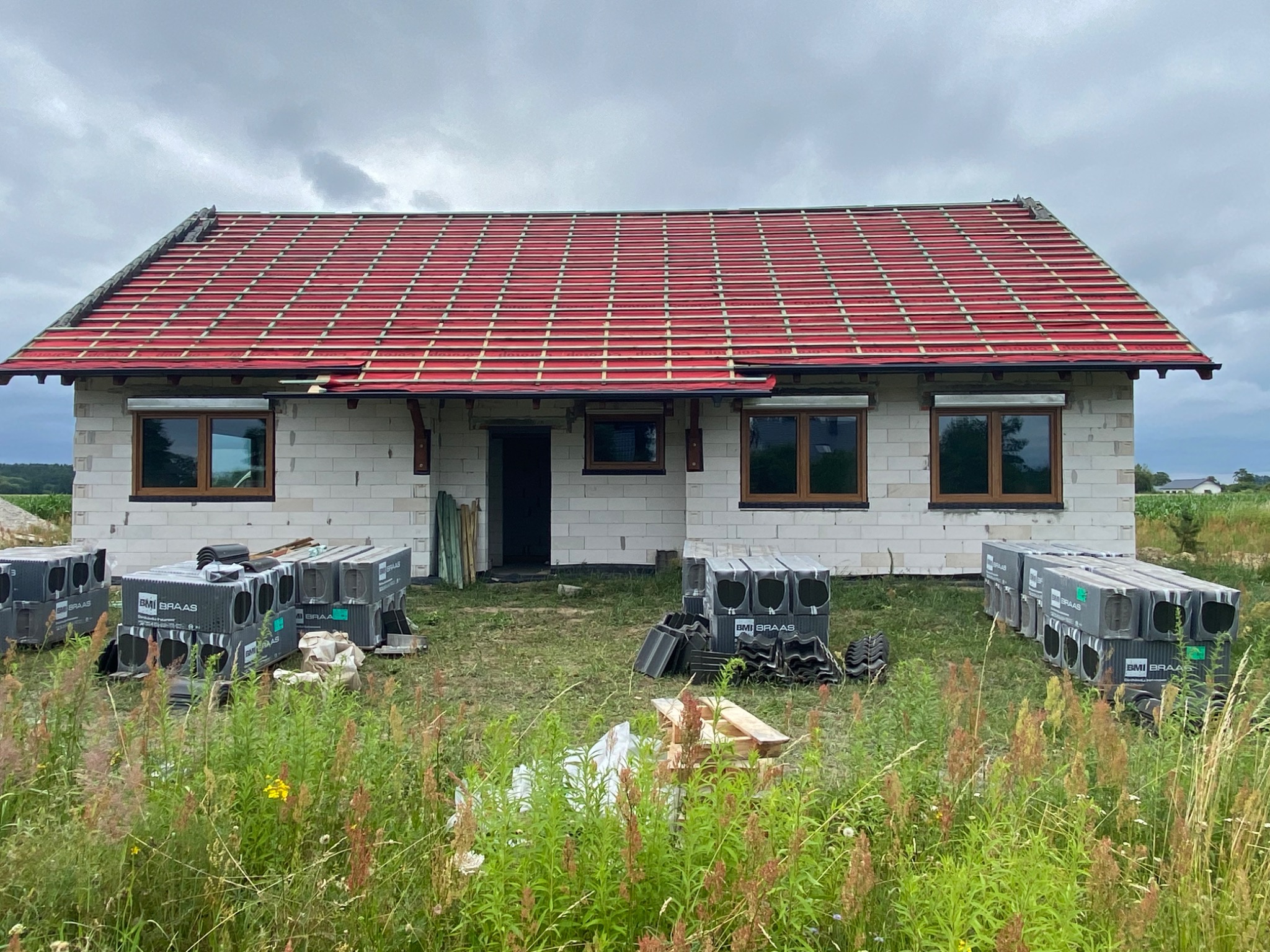 Niedokończony dom z czerwonym dachem, w oknach zamontowane rolety zewnętrzne, na trawniku ułożone palety z dachówkami BMI Braas.