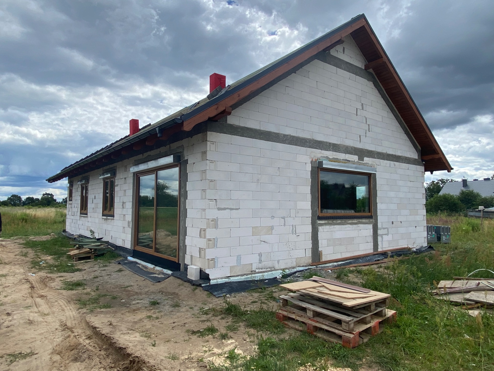 Surowy stan domu jednorodzinnego w trakcie budowy, widoczne okna z brązowymi ramami, ściany z białego betonu komórkowego, drewniana konstrukcja dachu i czerwone kominy, teren wokół domu częściowo...