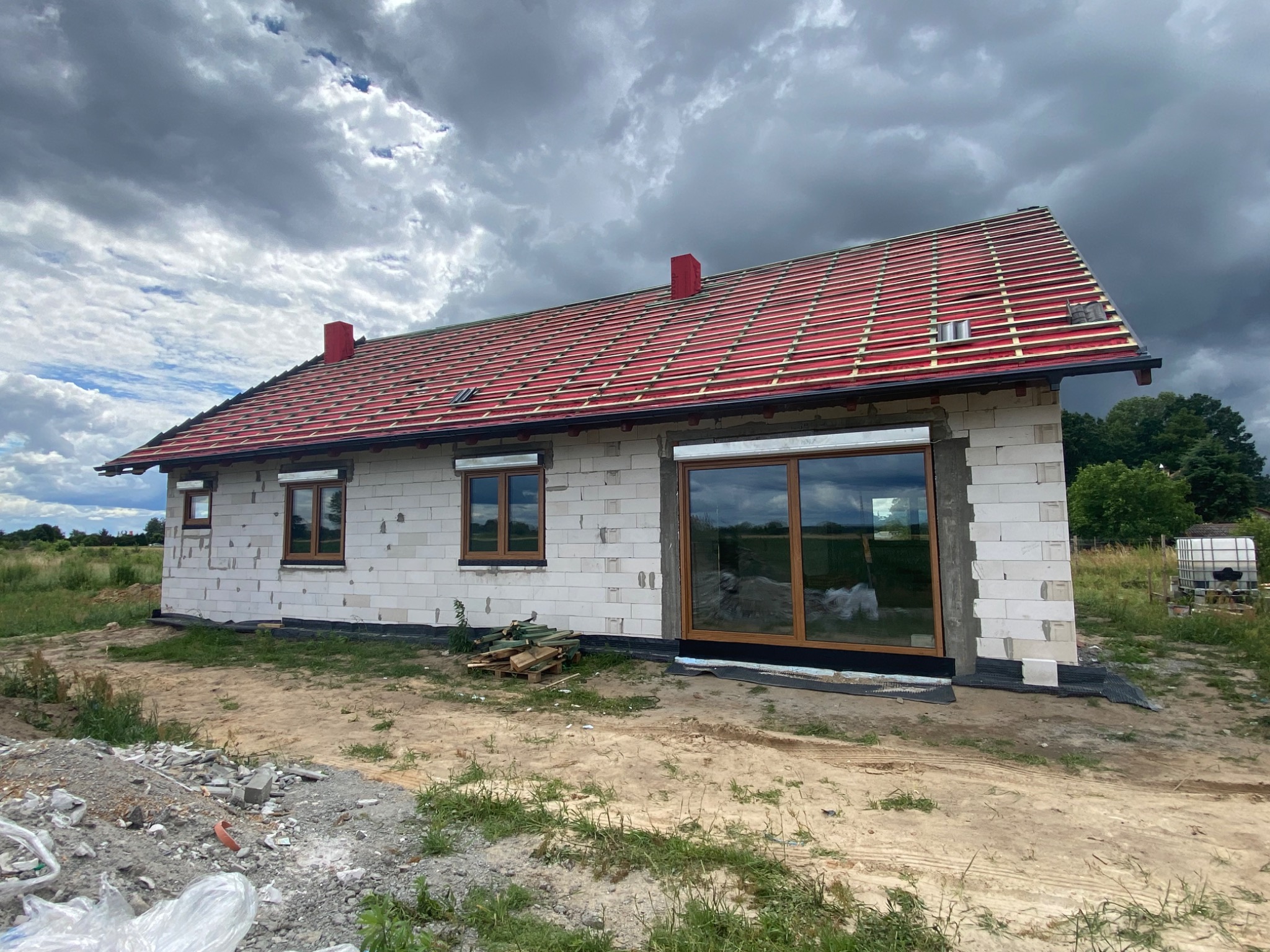 Surowy stan domu jednorodzinnego z czerwonym dachem i drewnianą stolarką okienną, widoczne niedokończone elewacje z białego bloczka i porozrzucane materiały budowlane.