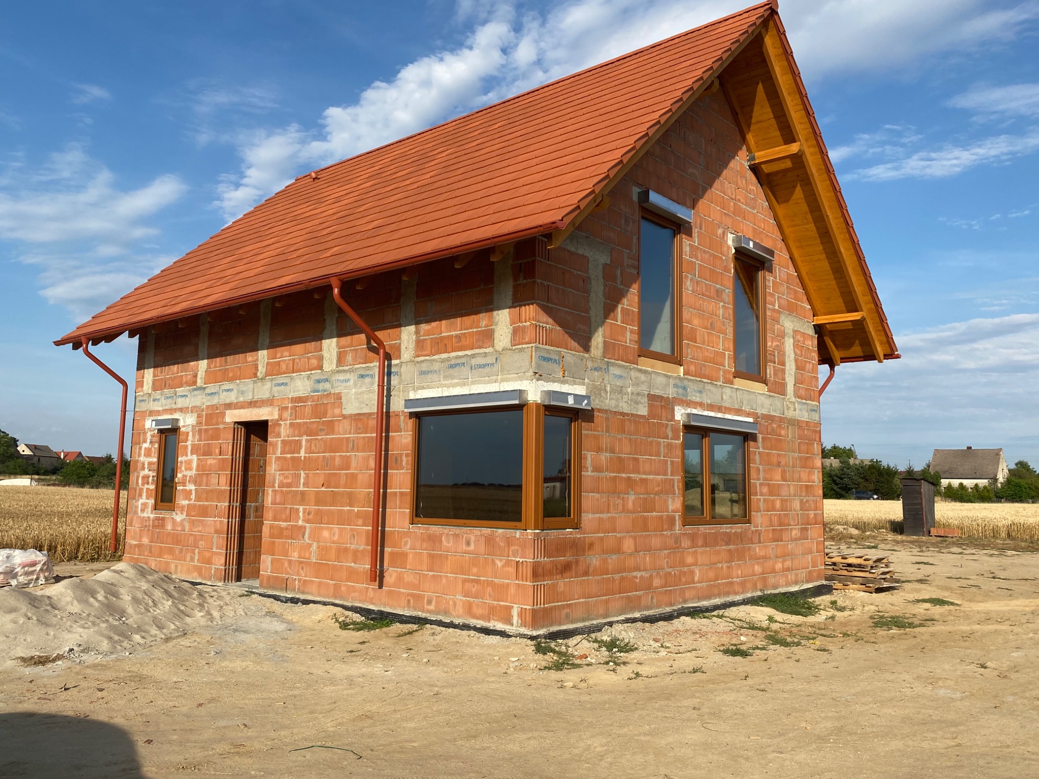 Surowy stan domu jednorodzinnego z czerwonej cegły, z zamontowanymi oknami PCV z brązowymi ramami i roletami zewnętrznymi, widok na pole zboża w tle.