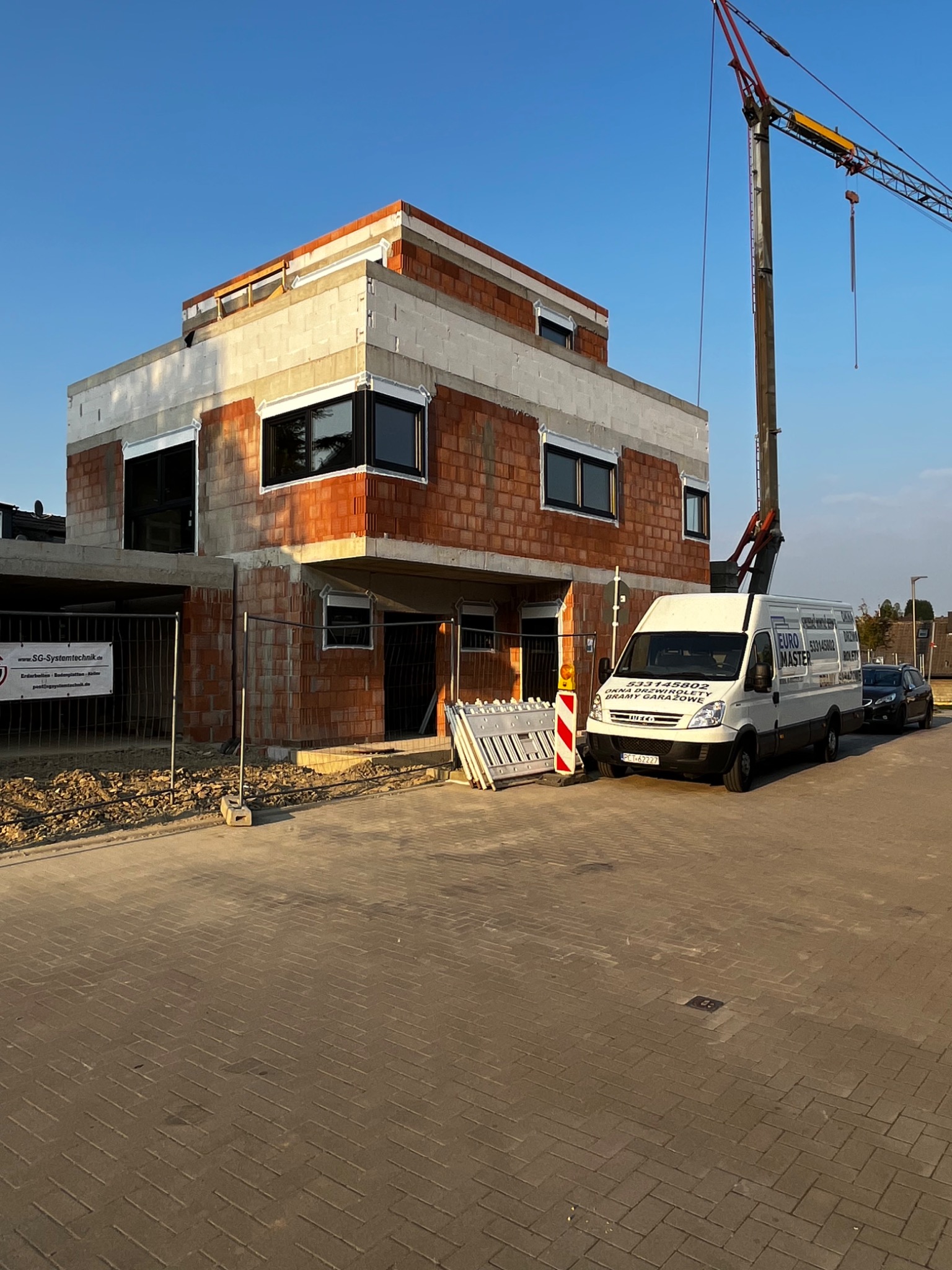 Budowa domu w trakcie wznoszenia, widoczne okna wstawione w surowy stan, obok zaparkowany biały bus z reklamą firmy budowlanej i dźwig budowlany.