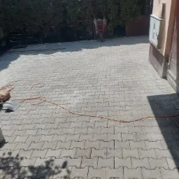 Ułożona szara kostka brukowa typu 'kość', widoczny fragment posesji, narzędzia pracy: łopata, przewód elektryczny, worek z materiałem, taczka w tle.