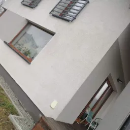 Jasnoszara elewacja domu jednorodzinnego z widocznymi oknami z brązowymi ramami i metalowymi kratami, drewniany taras z zielonymi krzesłami, obok trawnik z kamiennym obrzeżem.