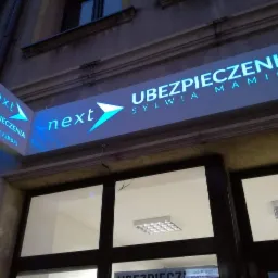 Oświetlony szyld firmy ubezpieczeniowej 'Next Ubezpieczenia Sylwia Mamica' nad wejściem do biura, z numerem telefonu widocznym na bocznej części szyldu.