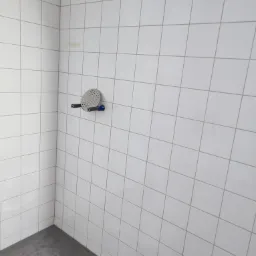 Pusta kabina prysznicowa z białymi kwadratowymi płytkami na ścianach, szarą podłogą i odpływem w rogu, widoczny uchwyt na słuchawkę prysznicową bez słuchawki, oraz fragment rury przy podłodze.