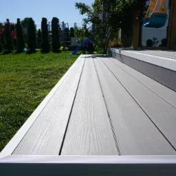 Szary taras z desek kompozytowych z aluminiowymi krawędziami, widok z poziomu trawnika, w tle zieleń ogrodu i huśtawka dla dzieci.