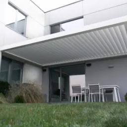 Nowoczesna, biała pergola z regulowanymi lamelami przy szarym budynku z dużymi oknami, widok z poziomu trawnika na meble ogrodowe.