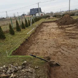 Przygotowany teren pod budowę domu z wytyczonym obrysem fundamentów za pomocą sznurka i palików, widoczny niwelator laserowy oraz pryzma ziemi.
