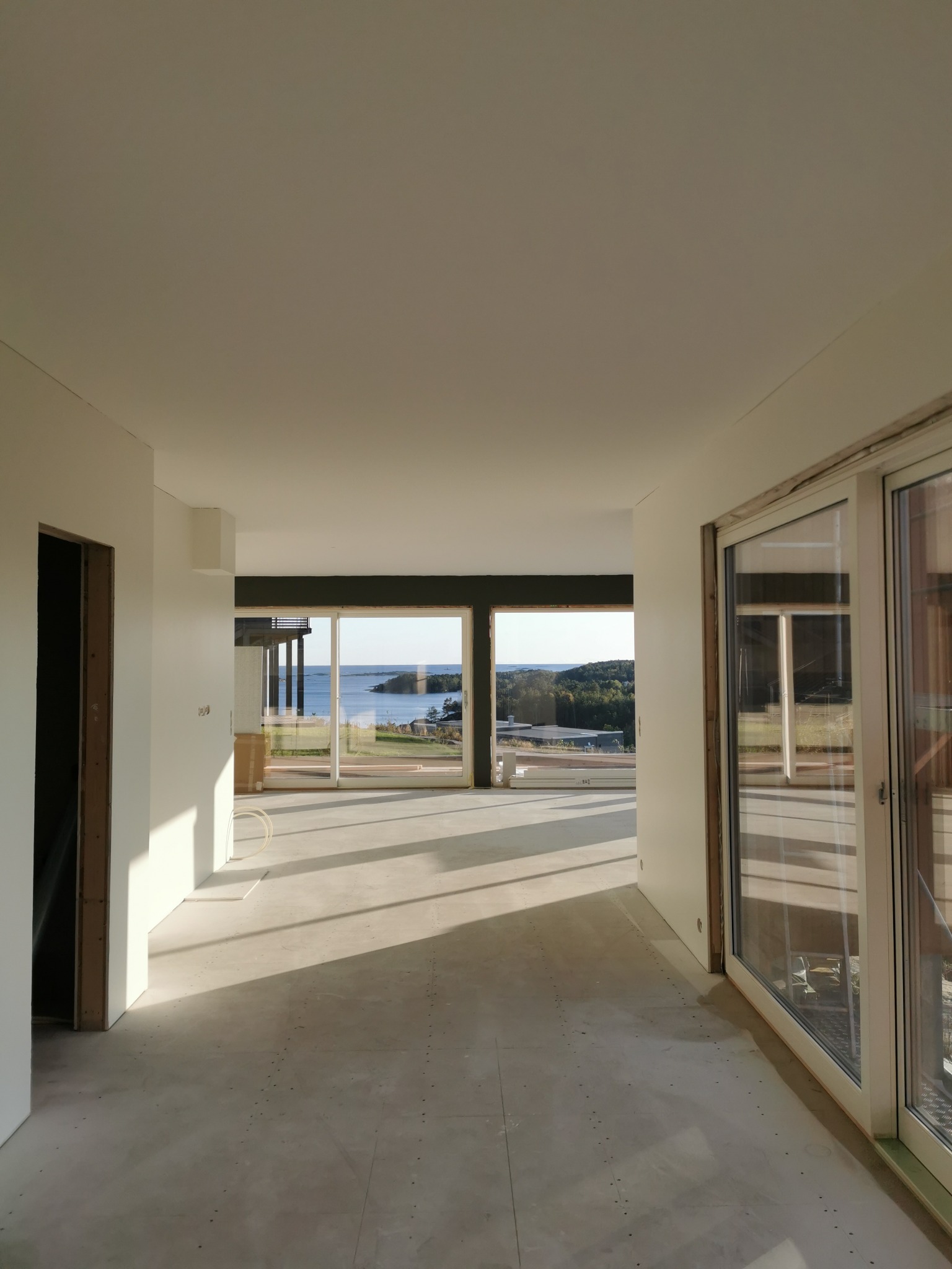 Wnętrze domu w trakcie wykańczania, świeżo pomalowane ściany na biało, duże okna z widokiem na morze, surowa podłoga, widoczne ślady po szpachlowaniu.