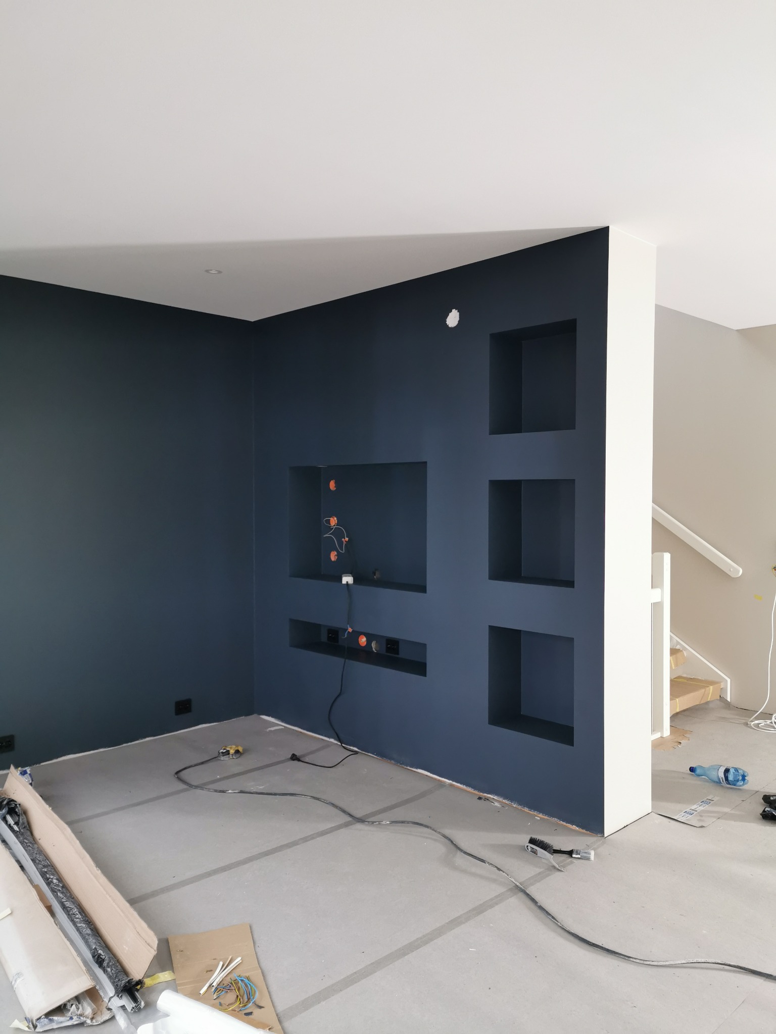 Świeżo pomalowana, ciemnoniebieska ściana z wnękami w salonie w trakcie wykańczania, widoczne kable i zabezpieczona podłoga.