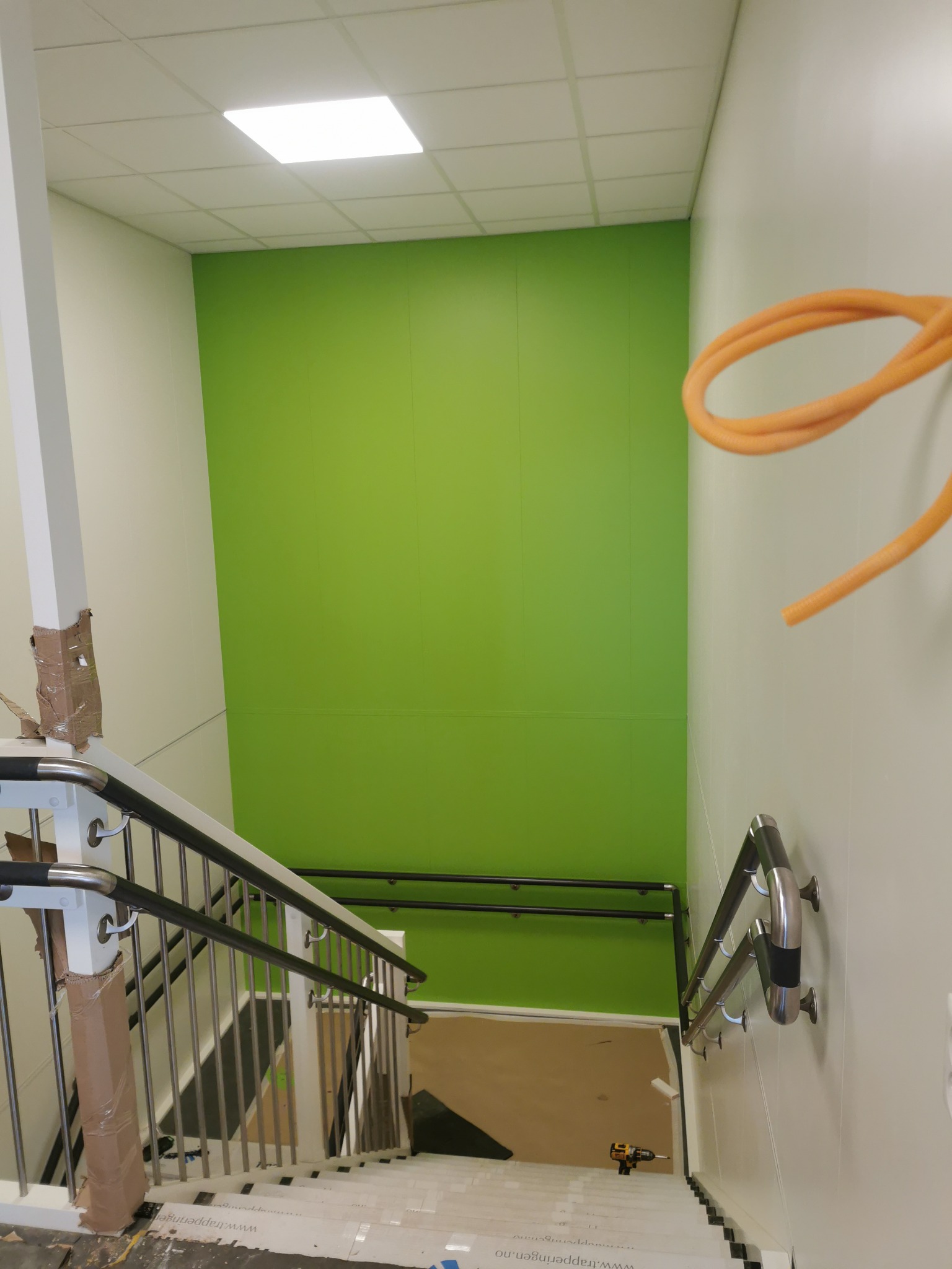 Klatka schodowa w biurze w trakcie wykończenia, zielona ściana, zabezpieczone schody, widoczne narzędzia i okablowanie.