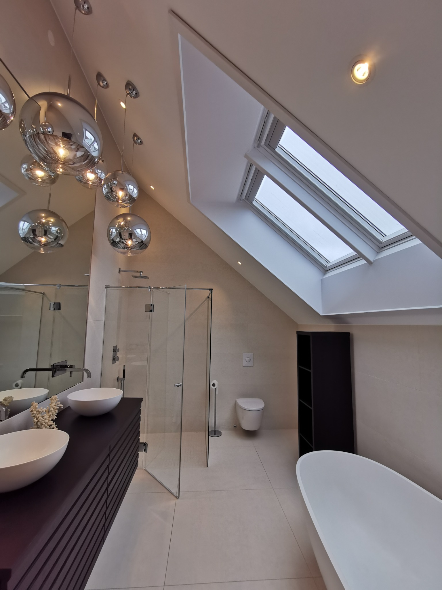 Nowoczesna łazienka na poddaszu z oknami dachowymi, kabiną prysznicową ze szklanymi drzwiami, umywalkami nablatowymi i dekoracyjnymi lampami kulami.