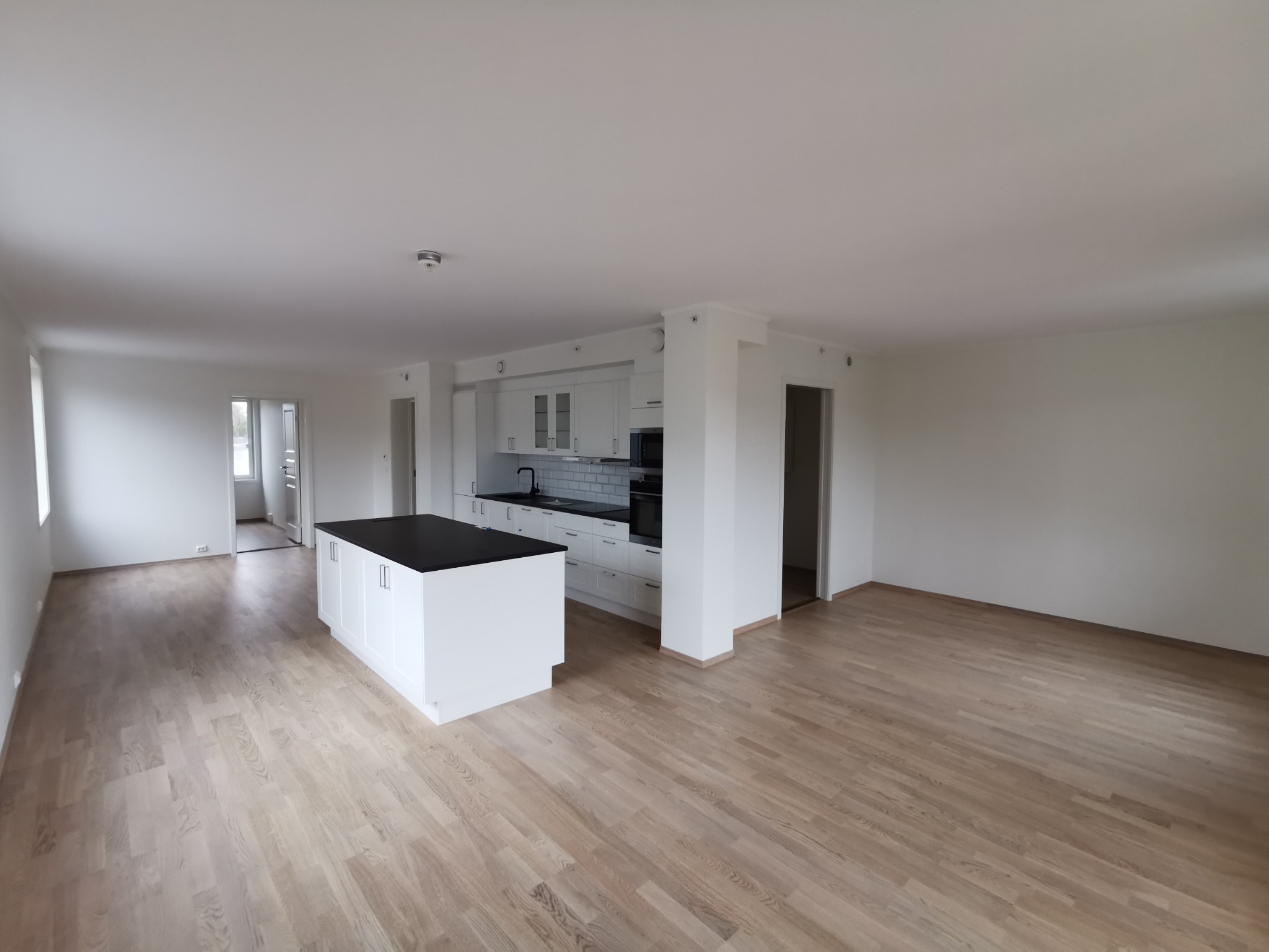 Jasne, przestronne wnętrze po remoncie: białe ściany, drewniana podłoga, kuchnia z wyspą. Widoczne gładkie powierzchnie ścian i sufitu.