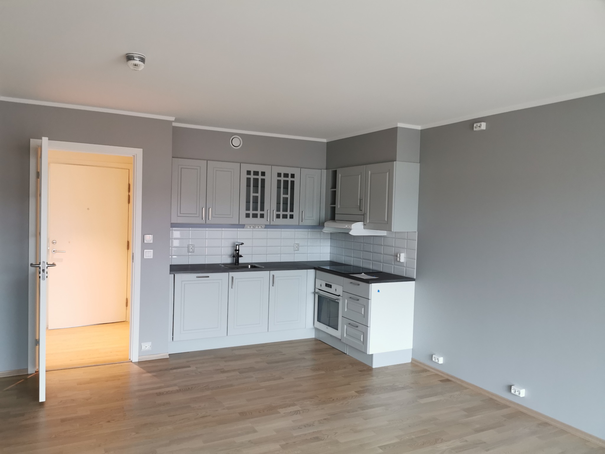 Wnętrze świeżo wykończonego mieszkania z szarą kuchnią i drewnianą podłogą, widoczne gniazdka elektryczne wzdłuż ściany i otwarte drzwi do innego pokoju.