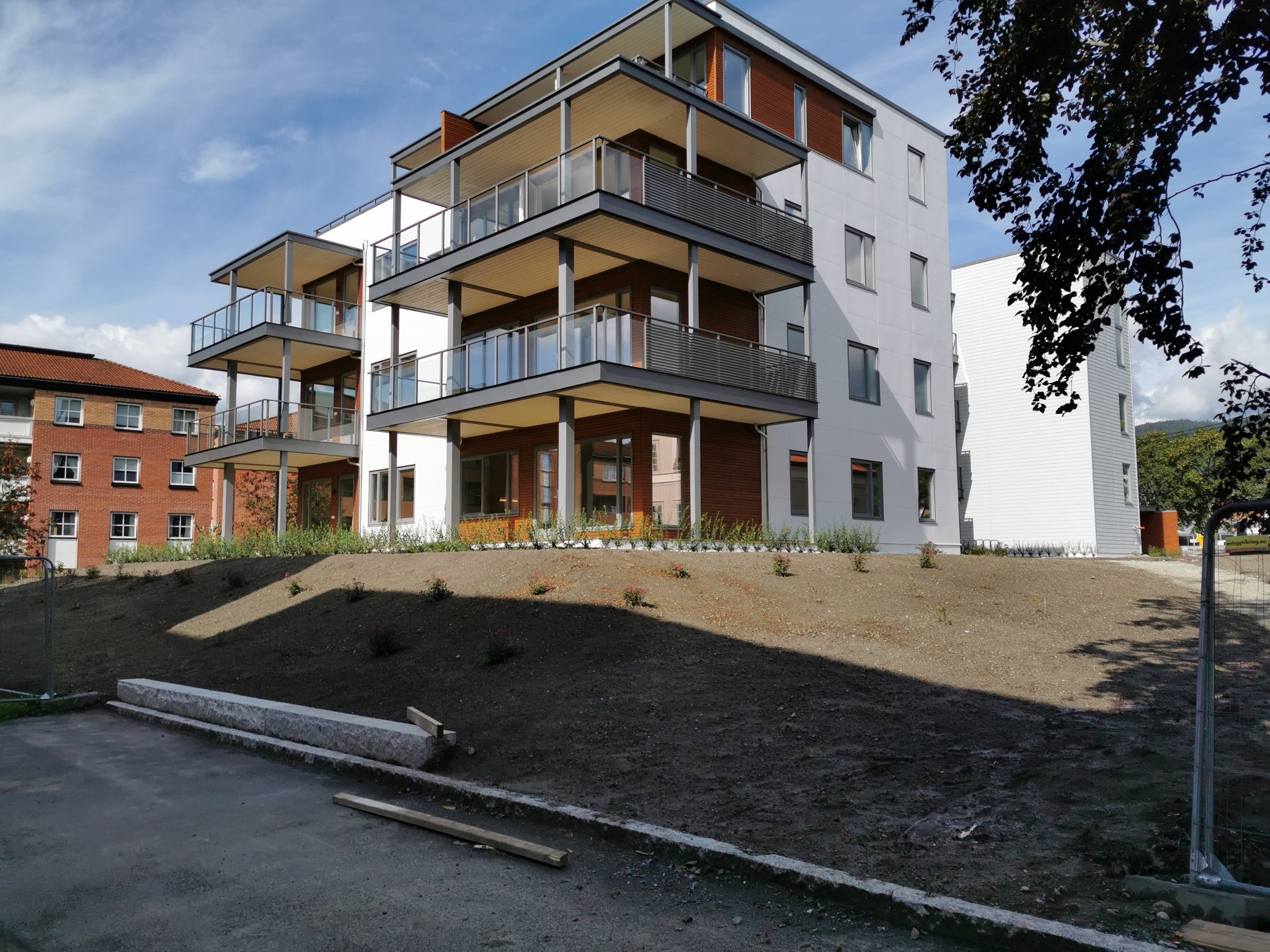 Nowoczesny budynek apartamentowy z balkonami, świeżo posadzona zieleń na zboczu, widok z dołu na błękitne niebo.