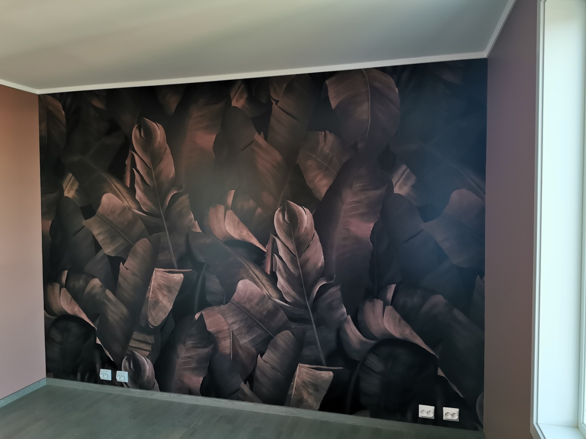 Ściana w pokoju z tapetą w motywie tropikalnych liści w odcieniach brązu i czerni, widoczne gniazdka elektryczne przy podłodze.