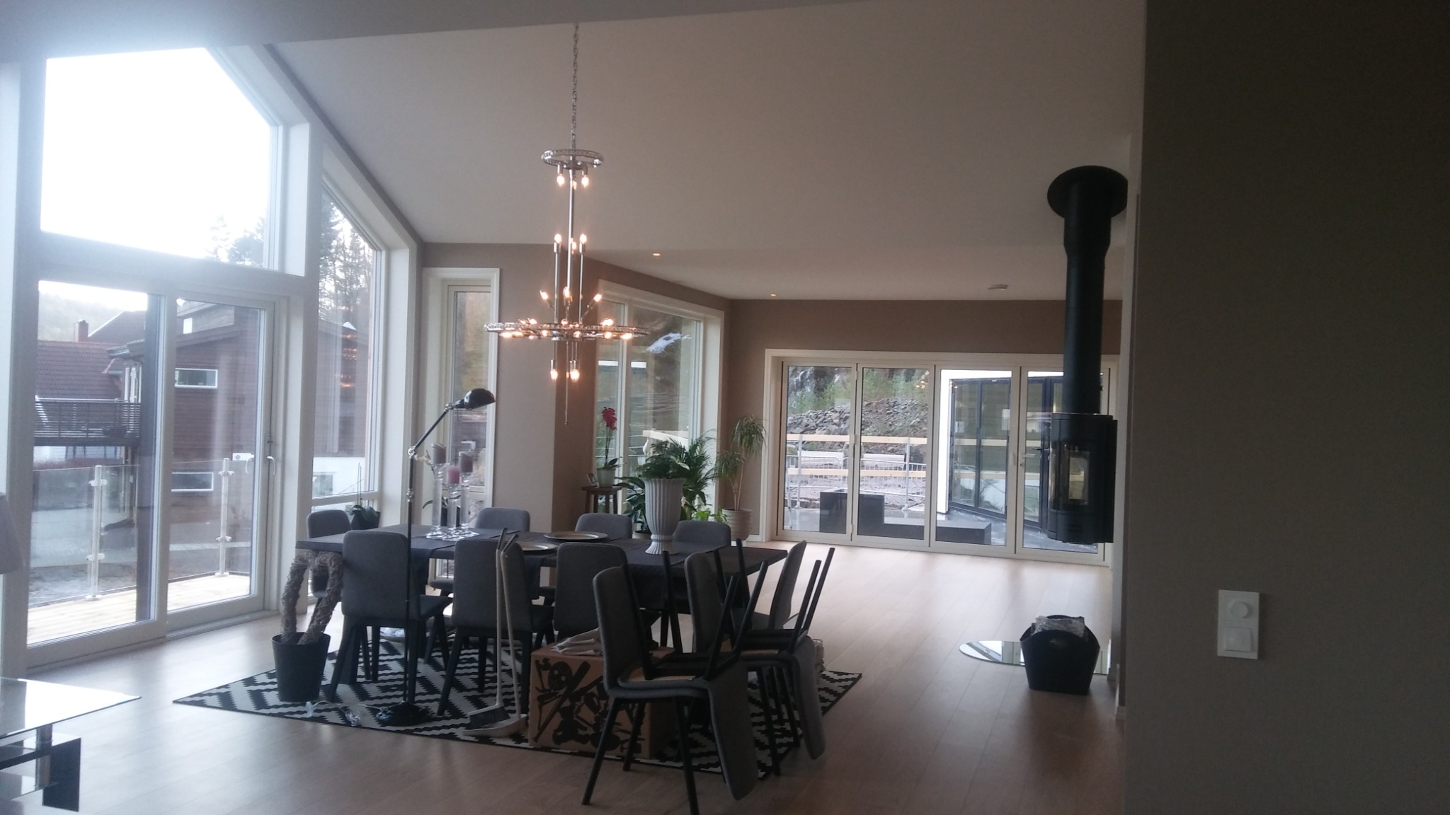 Jasne wnętrze domu z dużymi oknami, stołem z szarymi krzesłami, nowoczesnym żyrandolem i kominkiem wolnostojącym.