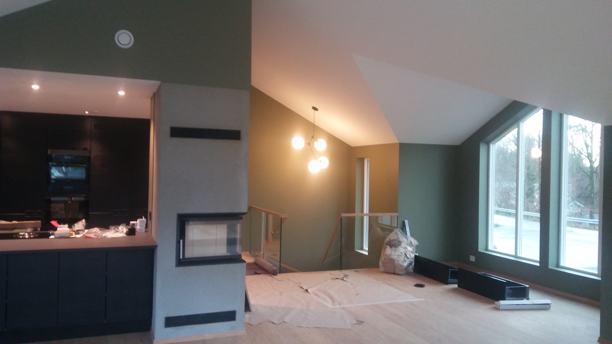 Wnętrze domu w trakcie prac wykończeniowych, z zielonymi ścianami, zabezpieczoną podłogą, nowoczesnym oświetleniem i widoczną kuchnią w tle.