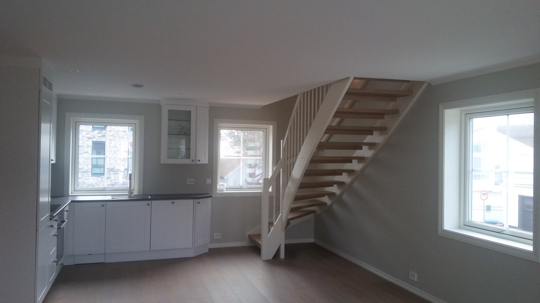 Jasne, świeżo pomalowane wnętrze z drewnianymi schodami, białymi meblami kuchennymi i widokiem z okna na ceglany budynek.