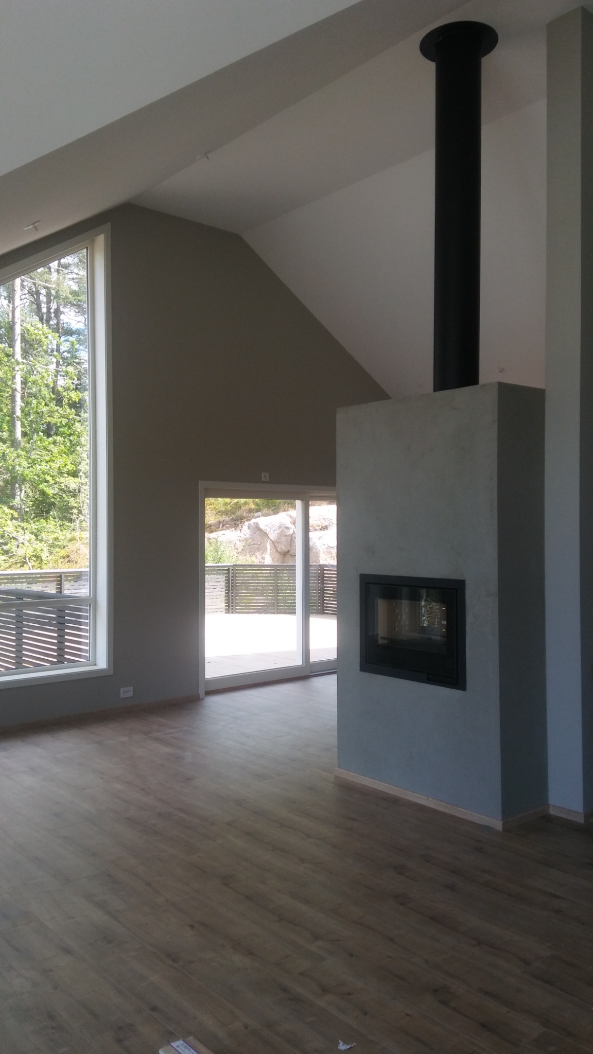 Wnętrze salonu z kominkiem o surowym wykończeniu, ściany w odcieniach szarości, duże okno z widokiem na zieleń i taras, drewniana podłoga.