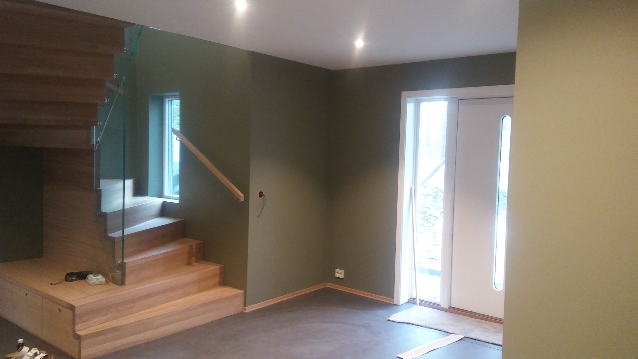 Wnętrze domu po remoncie: ściany w odcieniach zieleni, drewniane schody z szklaną balustradą, szara podłoga, białe drzwi wejściowe z oknami po bokach.