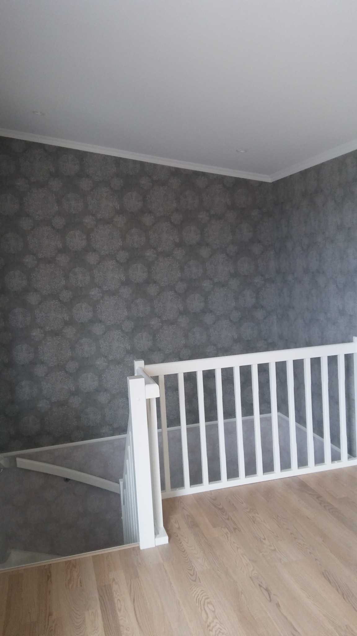 Wnętrze domu po remoncie: ściana pokryta szarą tapetą we wzory, biała balustrada schodów i drewniana podłoga.