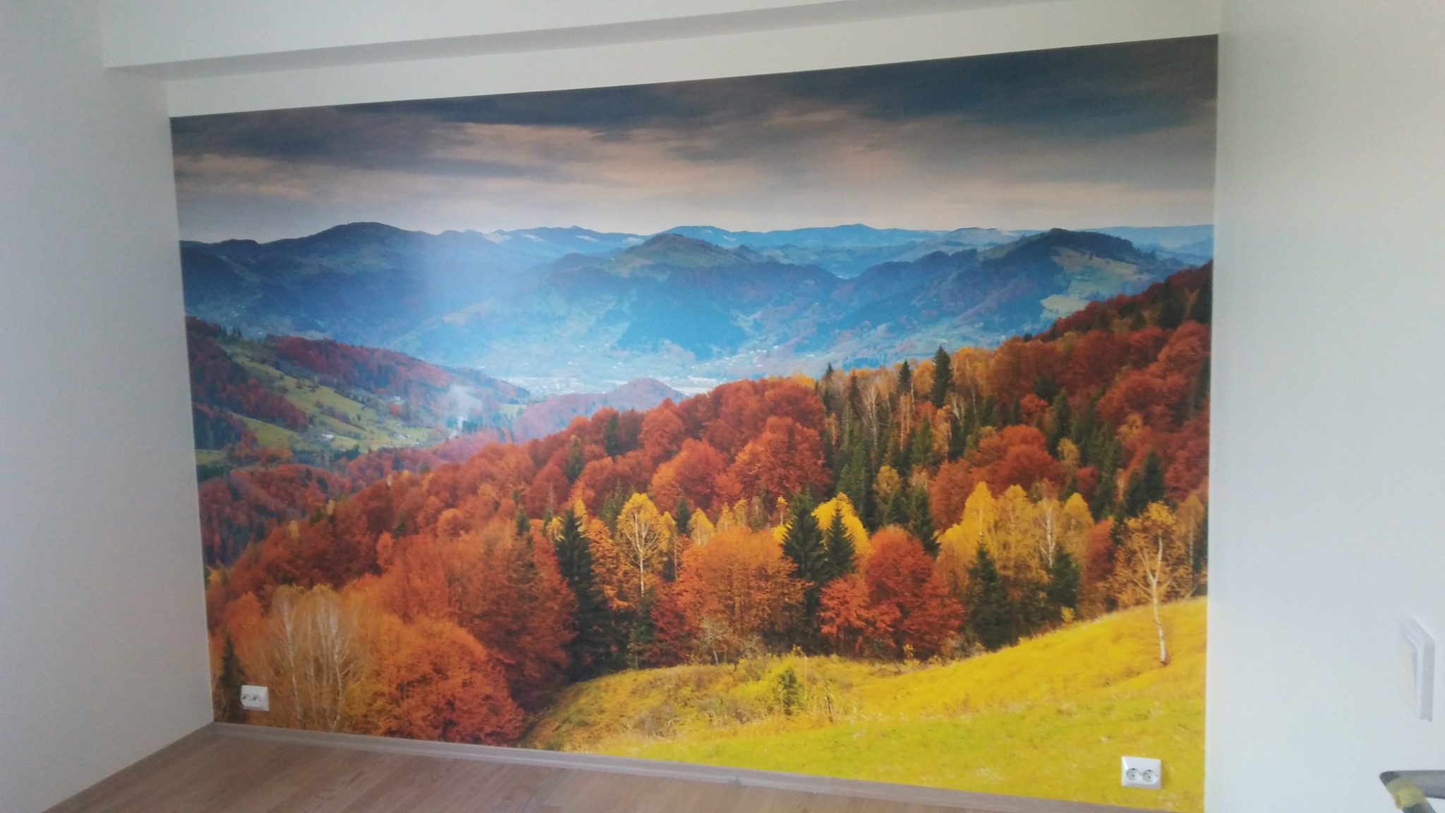 Fototapeta z jesiennym krajobrazem górskim na ścianie w jasnym pokoju, widoczne gniazdka elektryczne przy podłodze.