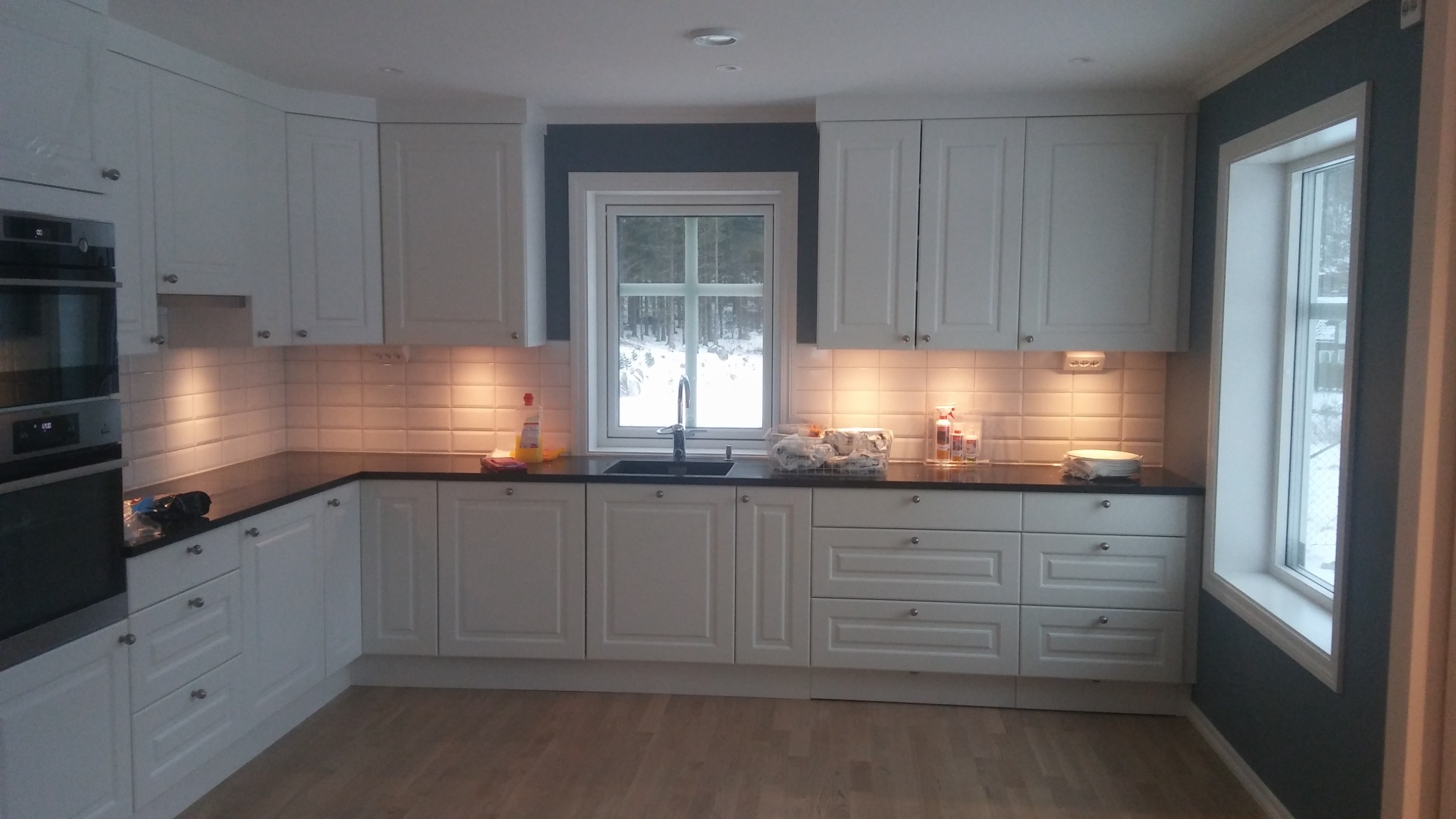 Wykończona kuchnia z białymi szafkami, ciemnym blatem i jasnymi płytkami na ścianie, widok na zaśnieżony krajobraz przez okno.