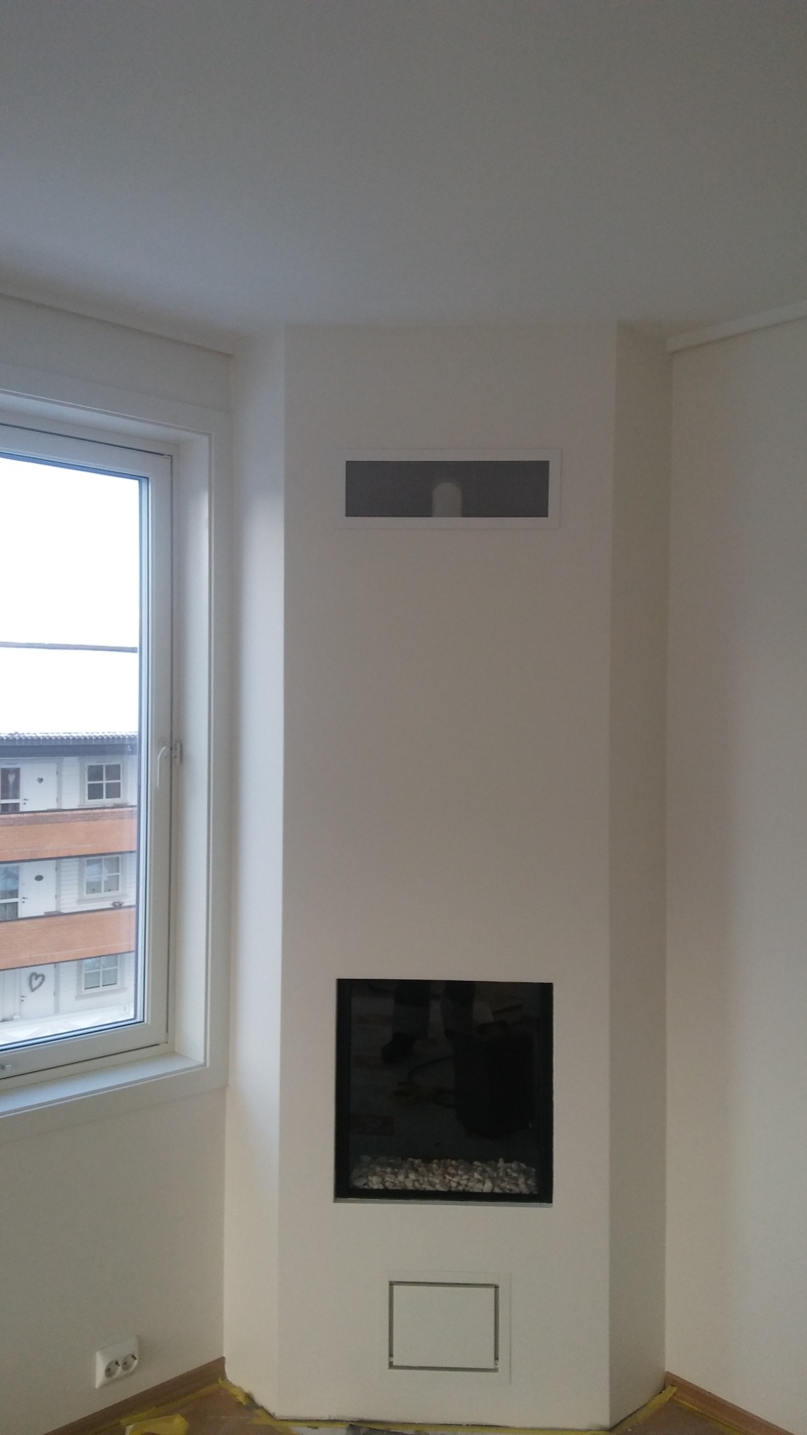 Wnętrze pokoju po remoncie, ściany pomalowane na biało, widoczny narożnik z oknem i wbudowany kominek elektryczny z kamieniami dekoracyjnymi, kratki wentylacyjne w ścianie.