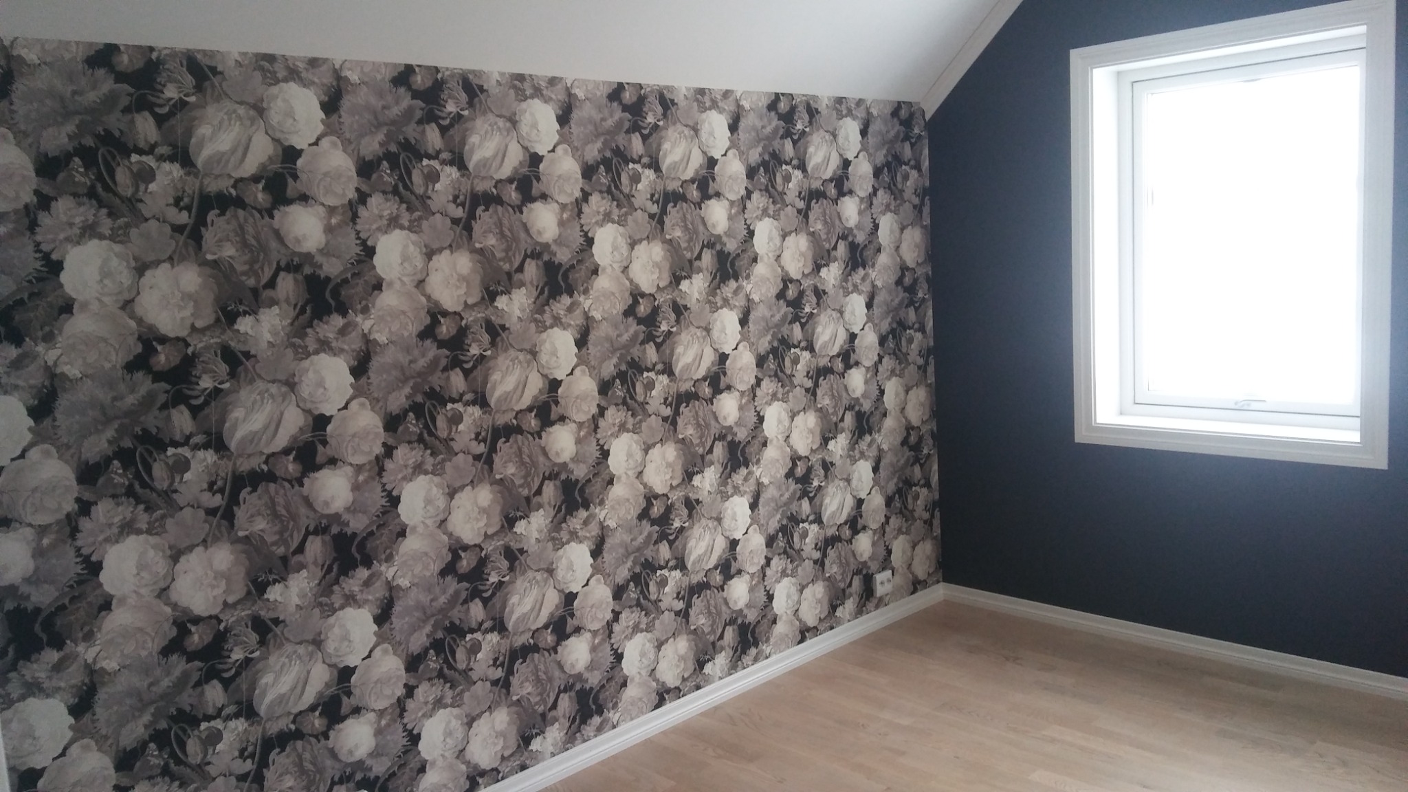 Pokój po tapetowaniu: Ściana pokryta wzorem kwiatowym w odcieniach szarości, sąsiadująca z ciemnoszarą ścianą z oknem, jasna drewniana podłoga i białe listwy przypodłogowe.