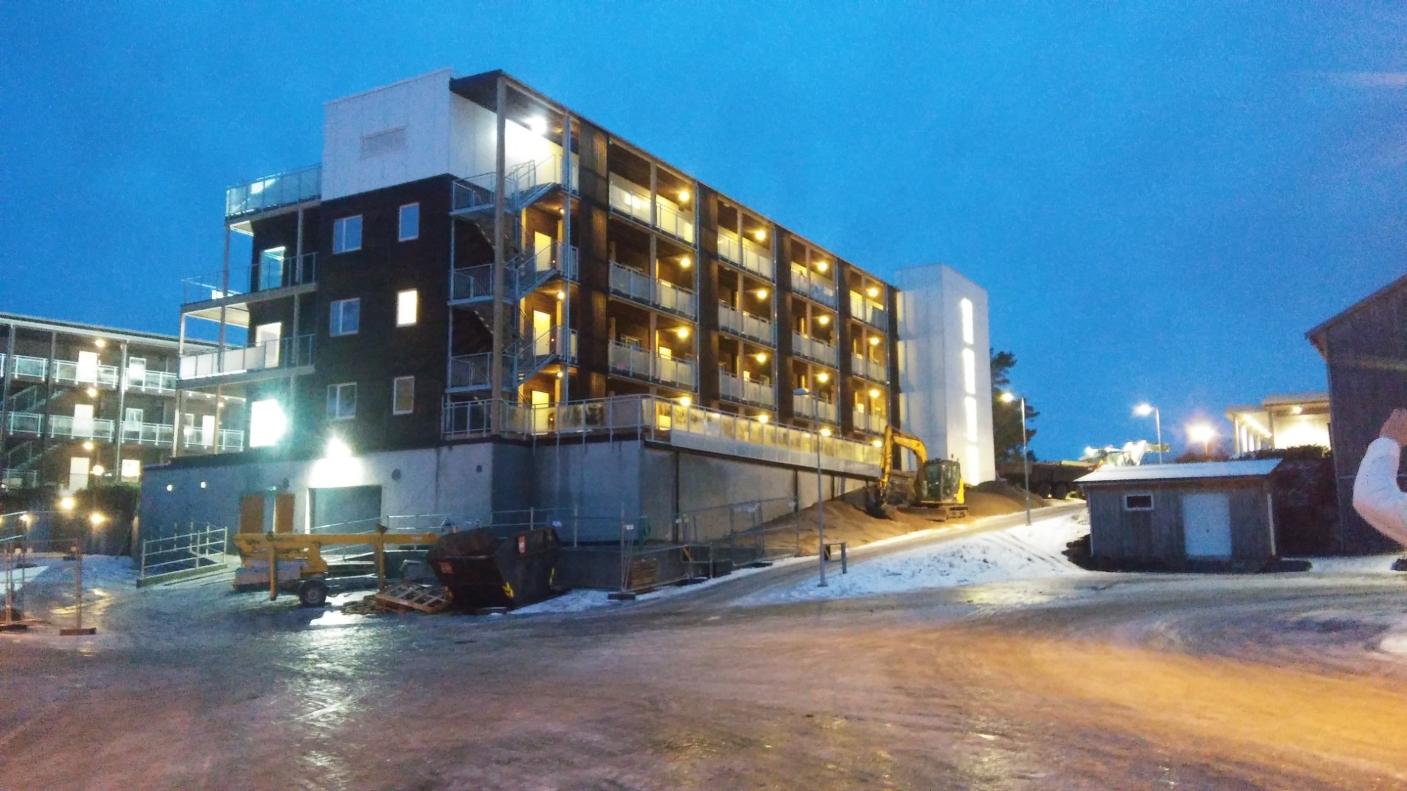 Wielopiętrowy budynek biurowy z balkonami i zewnętrznymi schodami ewakuacyjnymi, oświetlony w nocy, z widocznym sprzętem budowlanym i kontenerem na śmieci na placu budowy.
