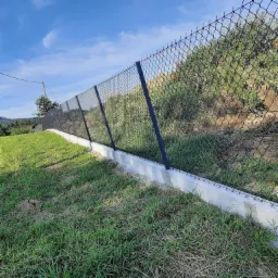 Ukończony montaż ogrodzenia z siatki na podmurówce betonowej, widok z dołu na zboczu, słupki w kolorze grafitowym.