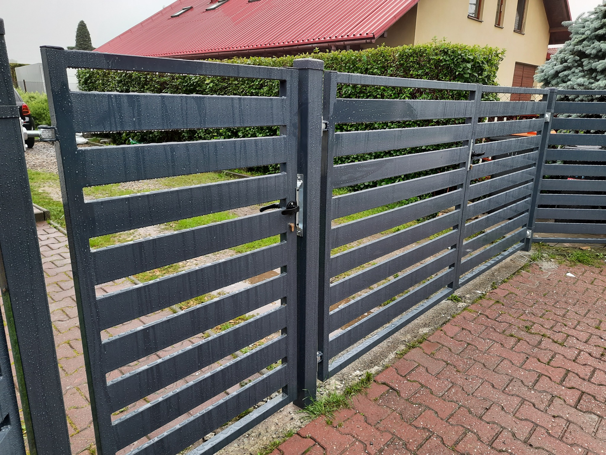 Nowoczesne, poziome ogrodzenie panelowe w kolorze grafitowym, z widocznymi kroplami deszczu, na tle domu z czerwoną dachówką i zielenią.