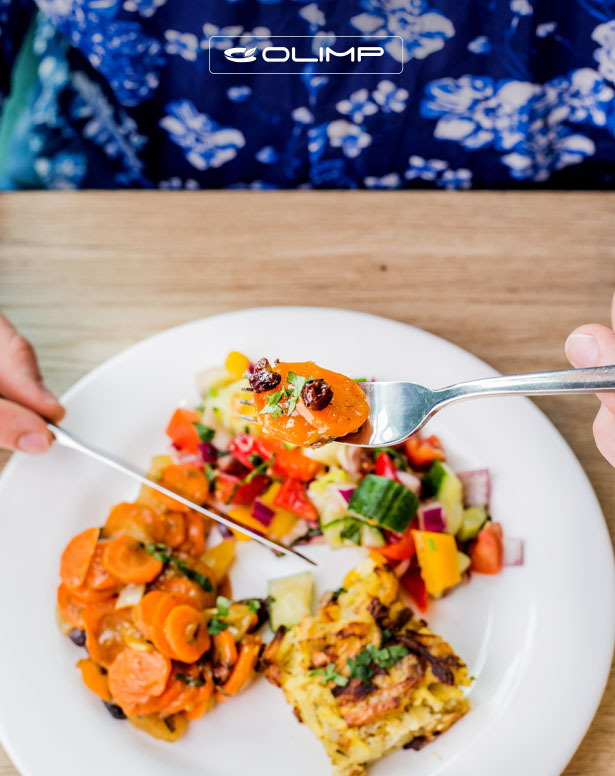 Osoba je posiłek z talerza, na którym znajdują się gotowane marchewki z czarnymi oliwkami, sałatka warzywna i zapiekanka ziemniaczana, widelec z kawałkiem marchewki w górze, na tle bluzki w kwiaty...