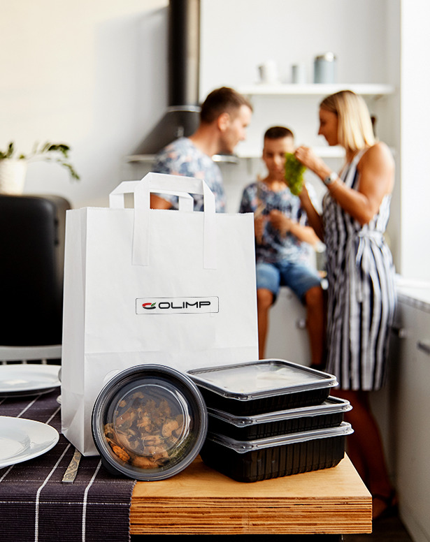 Posiłki w czarnych pojemnikach i torba papierowa z logo firmy Olimp na drewnianym blacie stołu z rodziną w tle, widok z góry z bliska.