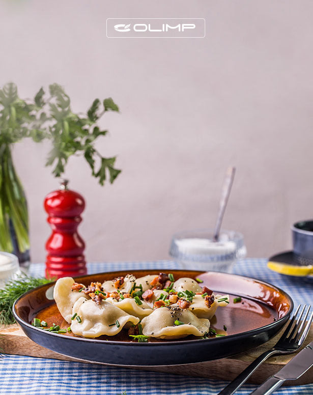 Apetyczne pierogi okraszone skwarkami i szczypiorkiem na brązowym talerzu, ułożone na drewnianej desce z niebieską kratą, w tle przyprawnik, zielenina i cukiernica.